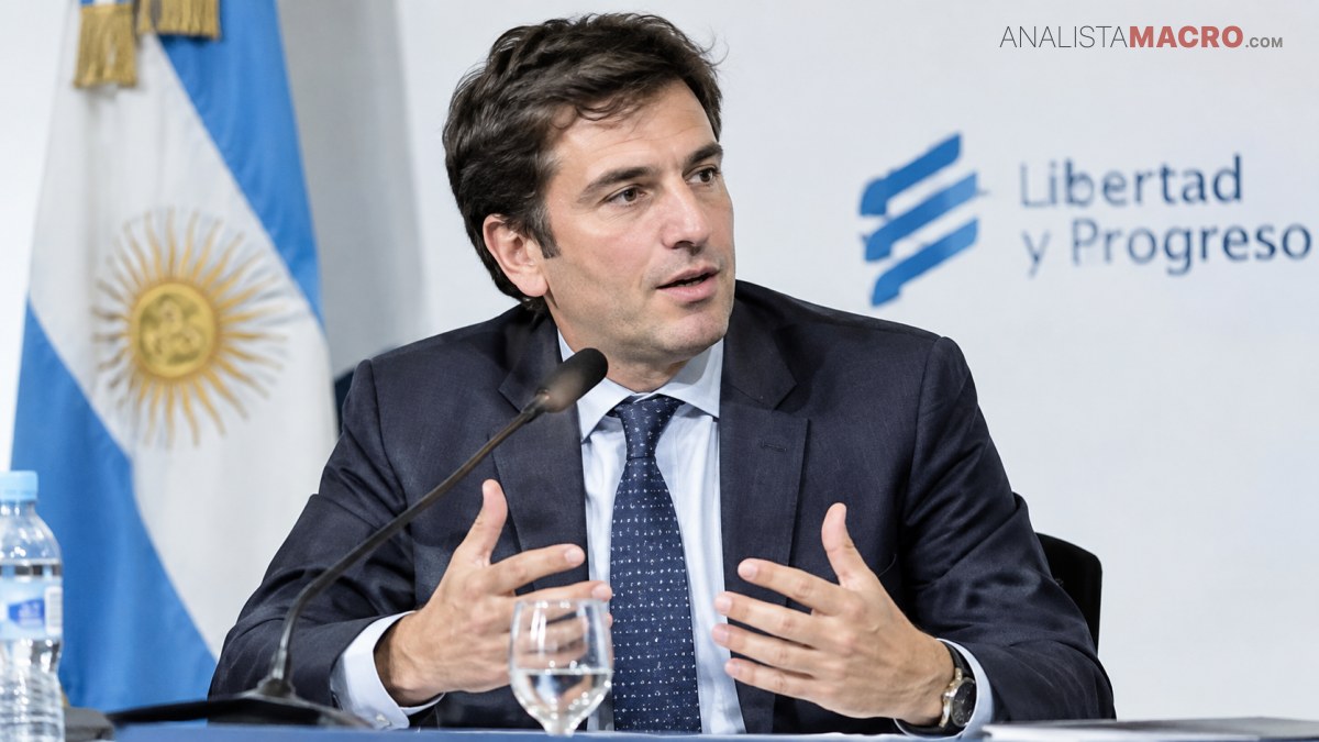 Hombre con traje y corbata habla ante un micrófono, gesticulando junto a una bandera argentina y un logotipo institucional.