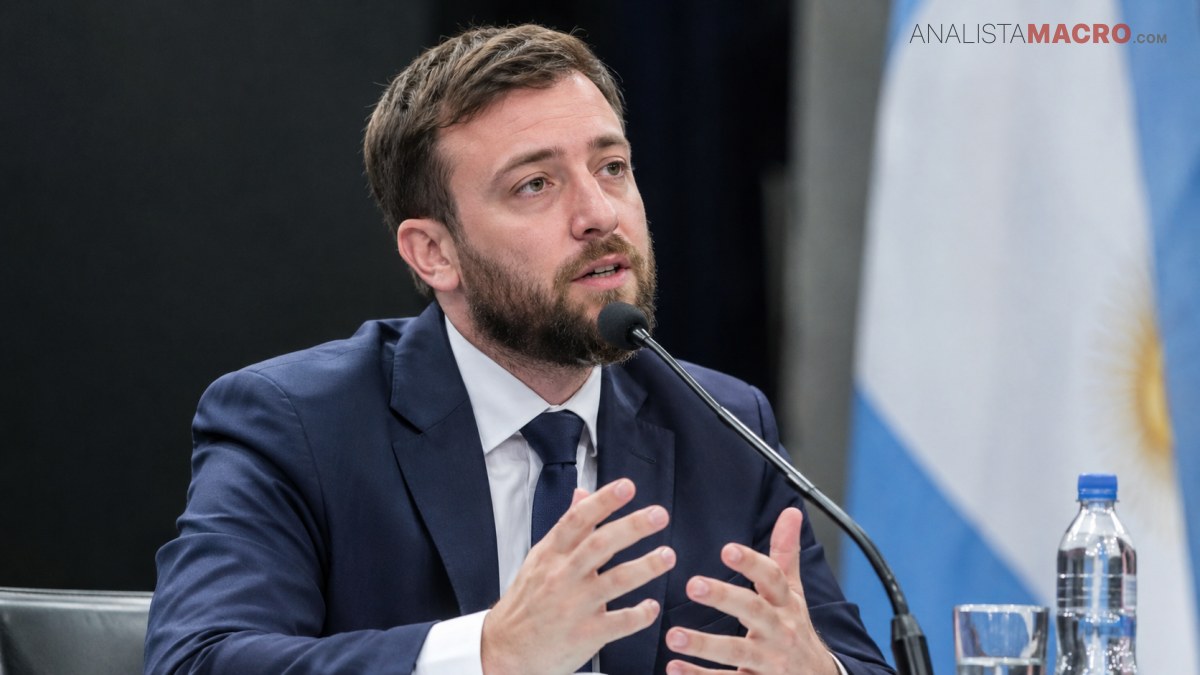 Hombre con traje azul y barba habla ante un micrófono, gesticulando con las manos junto a una bandera y botellas.