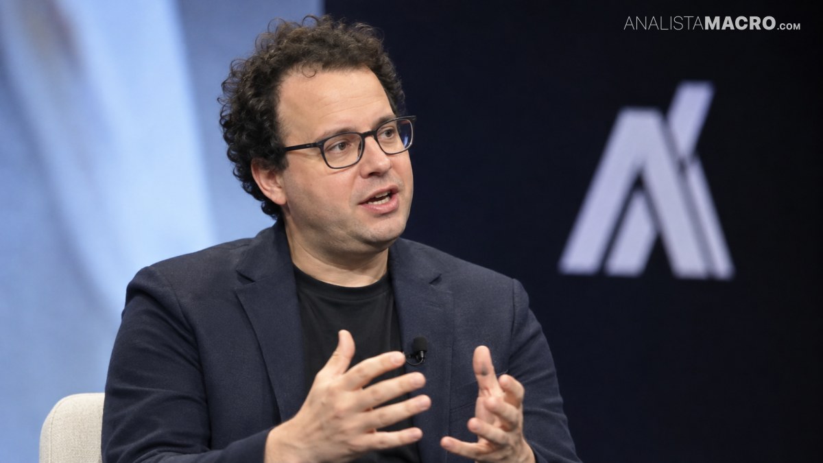Hombre con gafas, cabello rizado y chaqueta oscura gesticula mientras está sentado frente a un fondo con un logotipo.