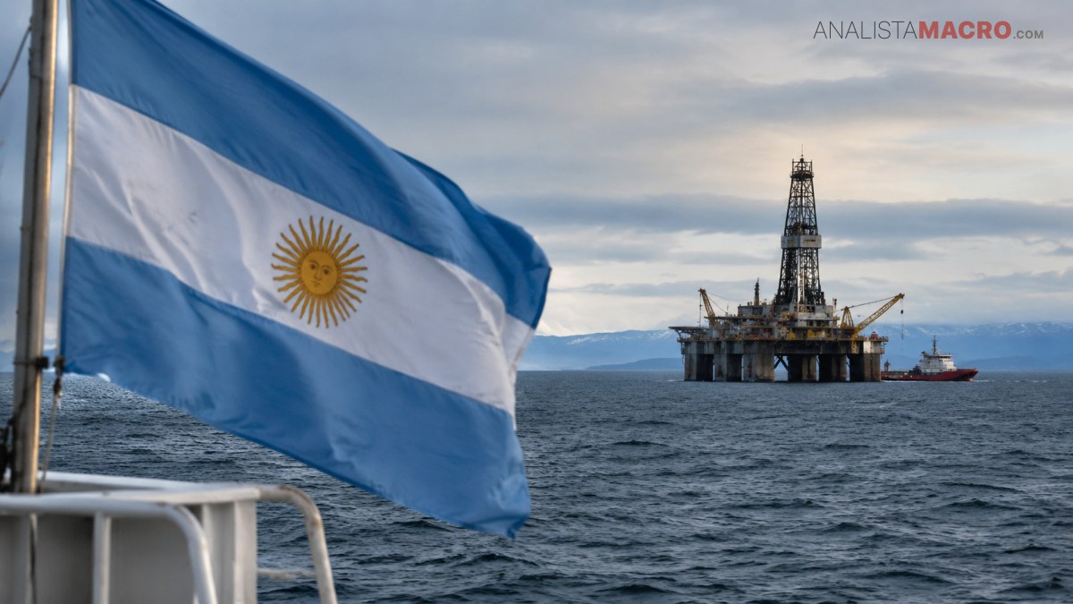 Una bandera argentina ondea en primer plano, frente a una plataforma petrolera marina y montañas bajo un cielo nublado.