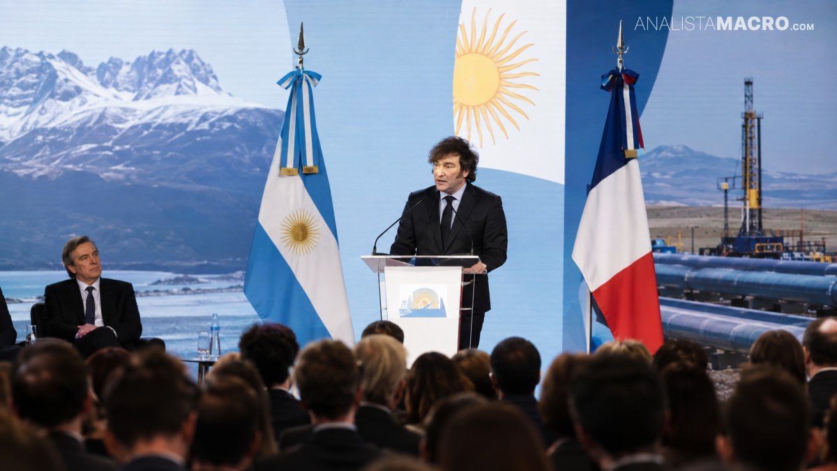 Un hombre habla desde un podio ante un público, acompañado por banderas argentinas y francesas y un paisaje montañoso.