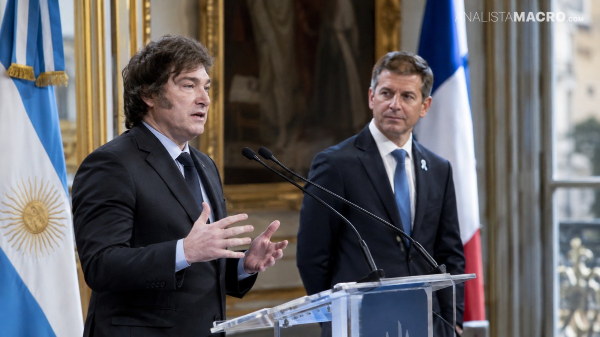 Dos hombres con traje están frente a micrófonos y un atril, con banderas argentinas al fondo.