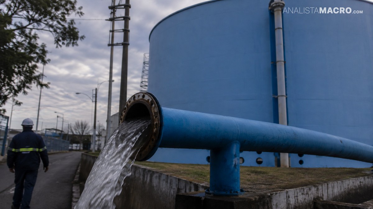Tubería azul descarga agua en un canal junto a un gran tanque industrial y una persona con casco camina al fondo.