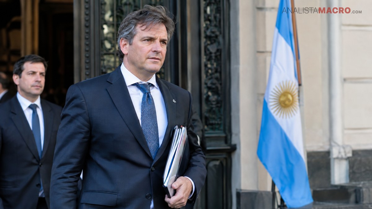 Hombre de traje oscuro sostiene documentos frente a un edificio, acompañado por otro hombre y una bandera argentina.