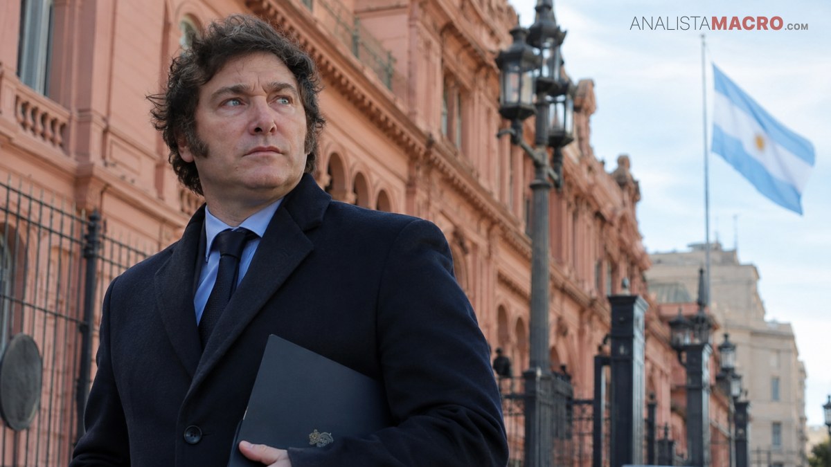 Hombre de traje oscuro sostiene una carpeta frente a un edificio rosado, con una bandera argentina ondeando al fondo.