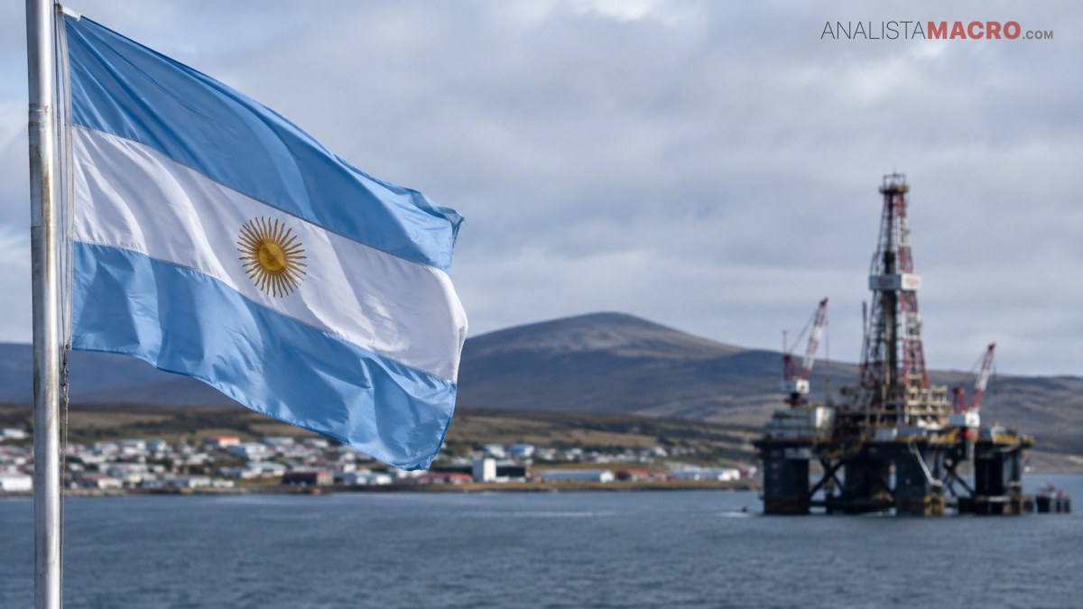 Bandera argentina ondeando frente a una plataforma petrolera marina, con montañas, viviendas costeras y cielo nublado al fondo.