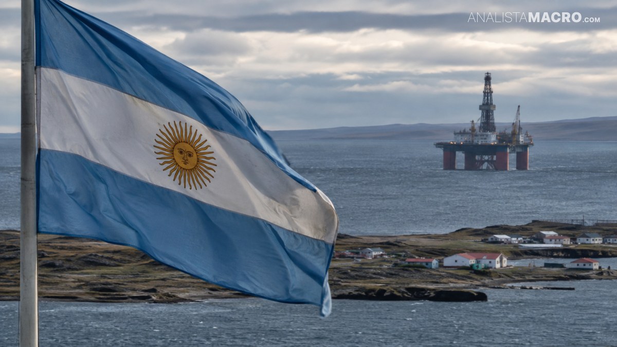 Bandera argentina ondeando frente al mar, con una plataforma petrolera y viviendas costeras al fondo.