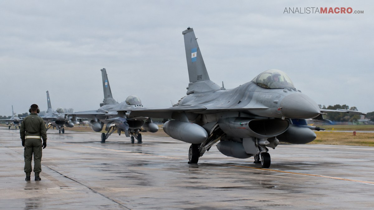 Varios aviones de combate alineados sobre una pista mojada, con una persona observándolos bajo cielo nublado.