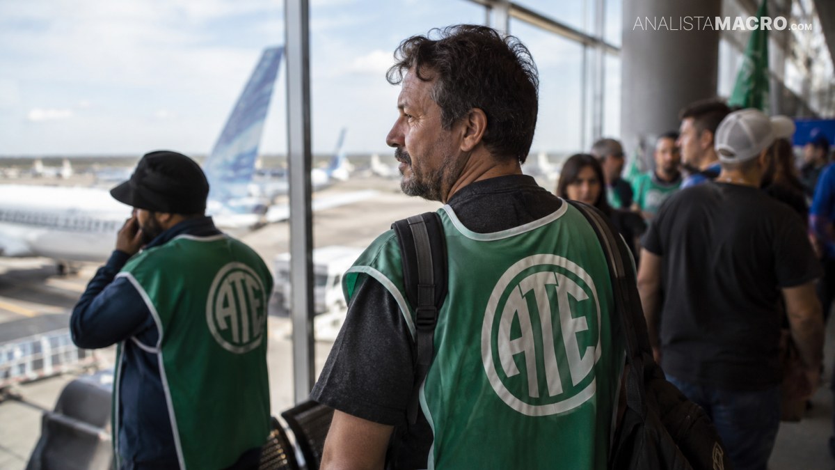 Dos hombres con chalecos verdes observan la pista de un aeropuerto, mientras varias personas esperan junto a los ventanales.