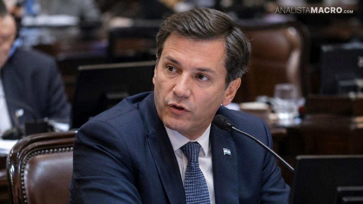 Hombre con traje azul y corbata habla ante un micrófono en una sala con escritorios y otras personas.