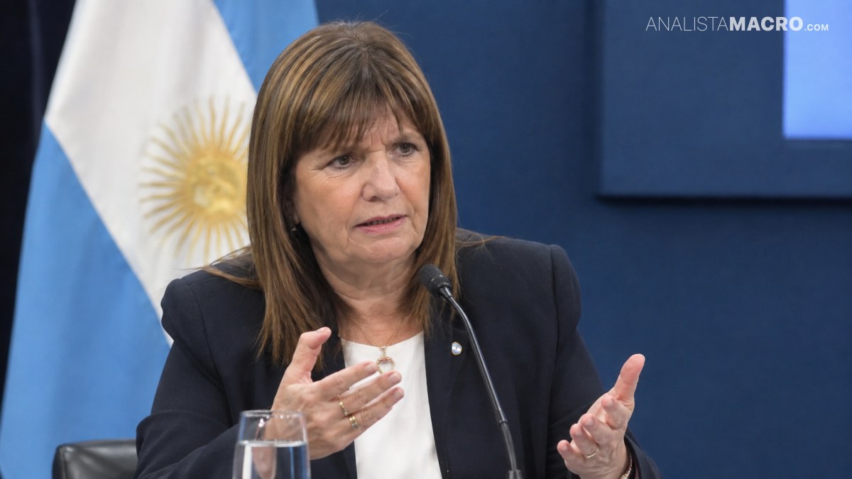 Mujer de cabello castaño habla ante un micrófono y gesticula con ambas manos, frente a una bandera argentina.