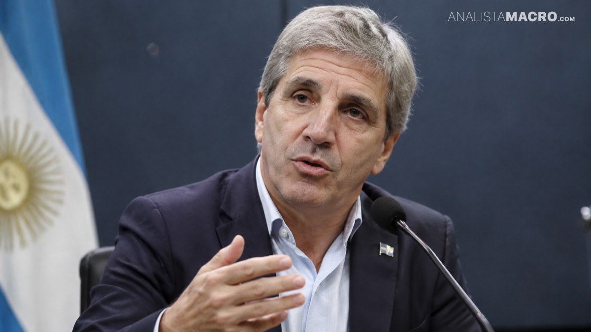 Hombre de cabello canoso habla ante un micrófono, gesticulando con una mano, mientras se ve parcialmente una bandera detrás.