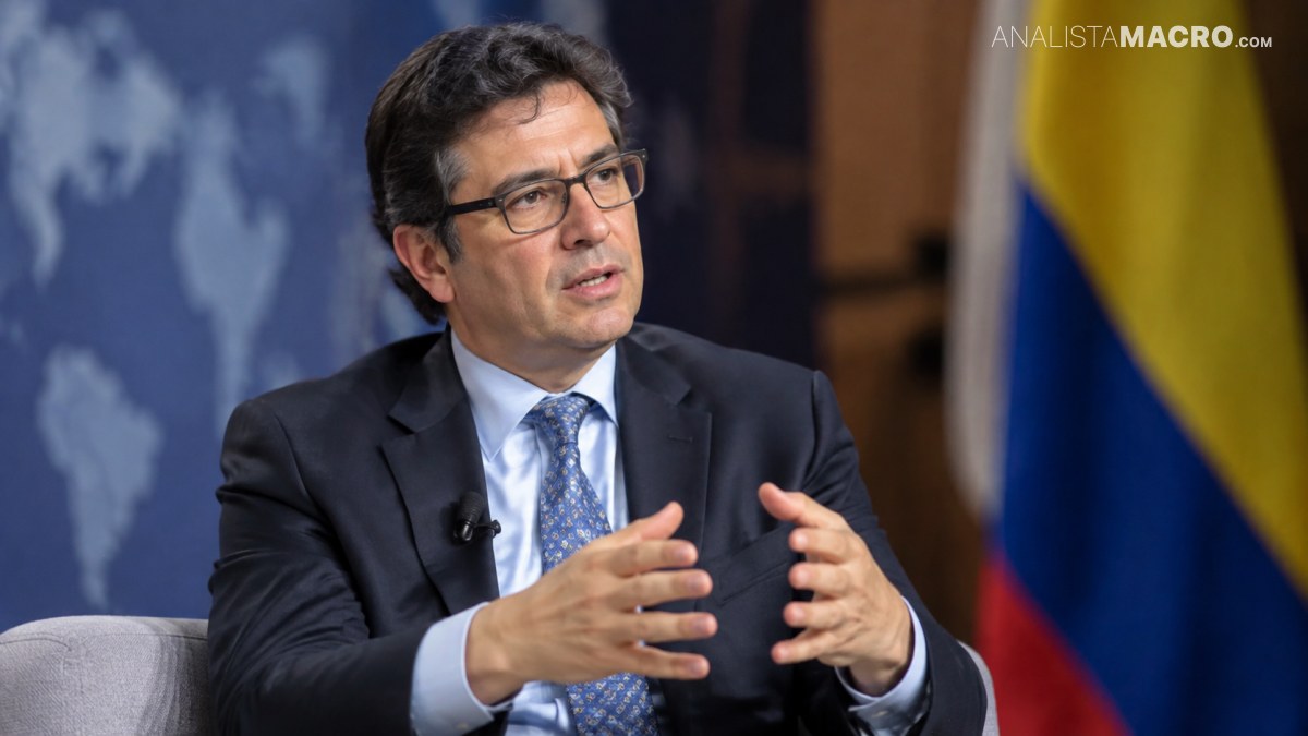 Hombre con gafas y traje oscuro gesticula con las manos, sentado frente a una bandera colombiana.