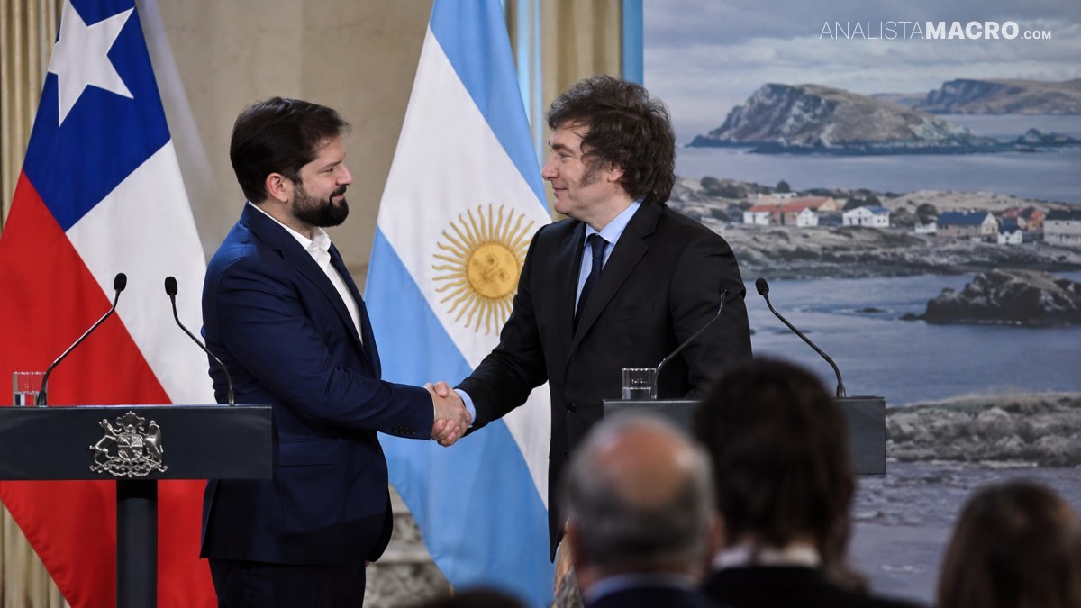 Dos hombres trajeados se estrechan la mano frente a banderas de Chile y Argentina, con un paisaje costero proyectado detrás.