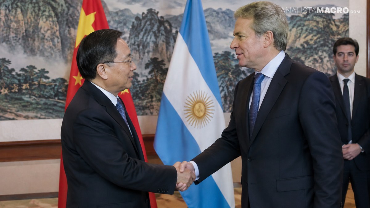 Dos hombres de traje se estrechan la mano frente a las banderas de China y Argentina, con un tercer hombre detrás.