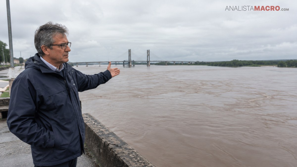 Hombre con gafas y chaqueta oscura señala un río crecido bajo un cielo nublado, junto a un muelle.