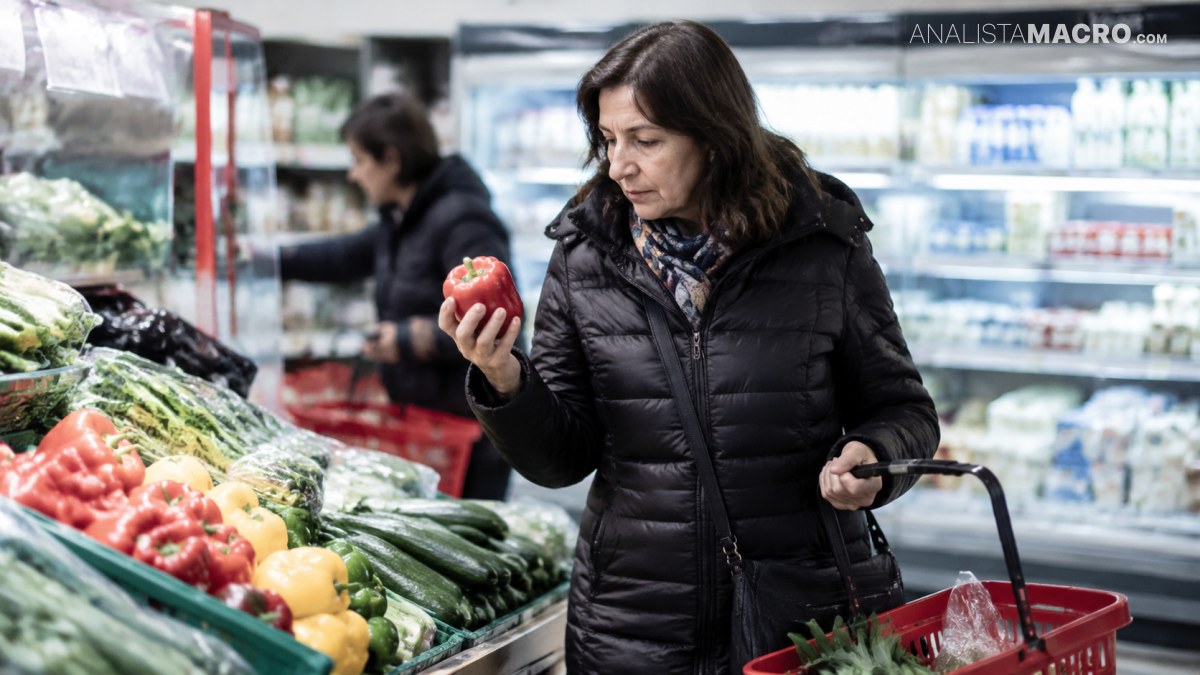Mujer con chaqueta negra sostiene un tomate junto a un carrito rojo, rodeada de verduras en un supermercado.