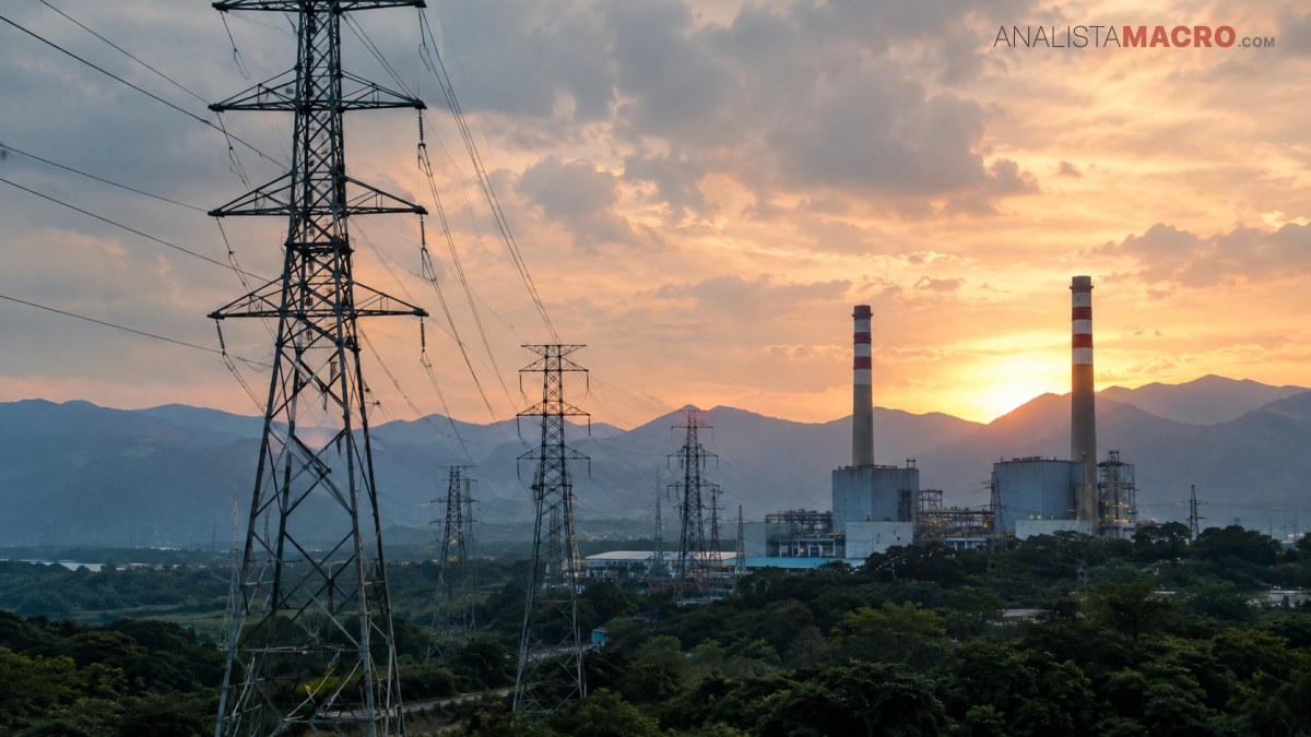 Torres eléctricas y una central con chimeneas se ven al atardecer, con montañas al fondo y nubes rosadas.