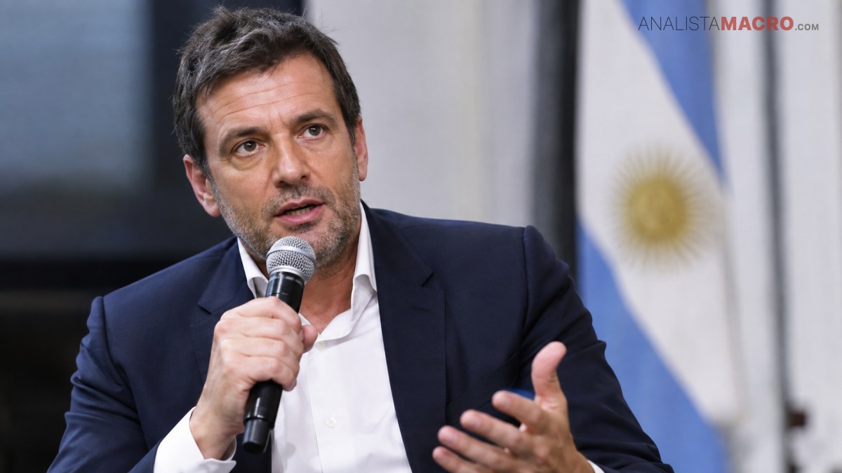 Hombre con traje oscuro y camisa blanca sostiene un micrófono y gesticula frente a una bandera argentina.