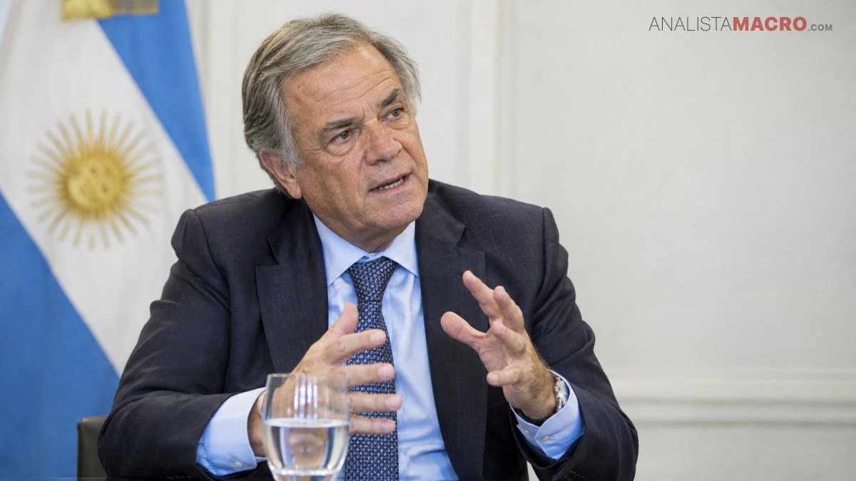 Hombre de cabello canoso y traje oscuro gesticula mientras habla, sentado frente a una bandera argentina.