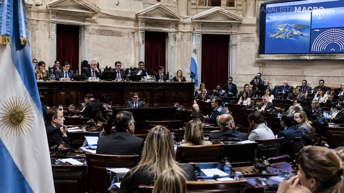 Varias personas participan en una sesión legislativa, sentadas frente a un estrado y rodeadas de escritorios, documentos y banderas.
