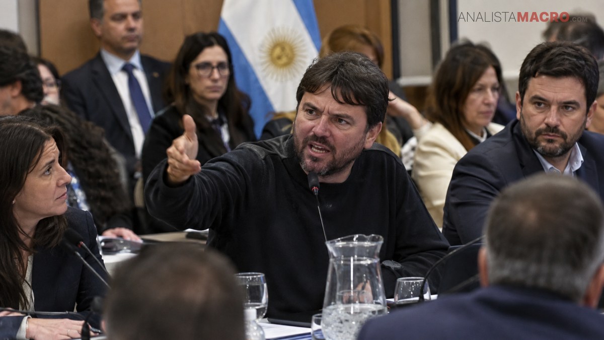 Un hombre gesticula mientras habla ante un micrófono, rodeado de varias personas sentadas alrededor de una mesa.
