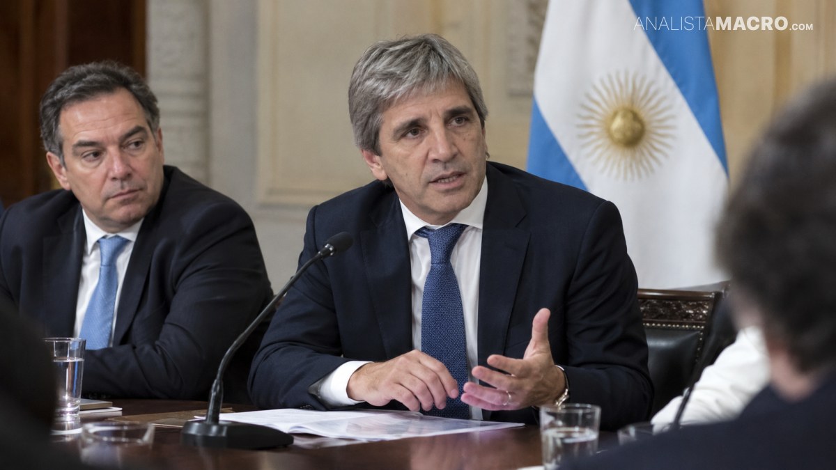 Dos hombres sentados en una mesa; el del centro habla y gesticula, mientras una bandera argentina aparece al fondo.