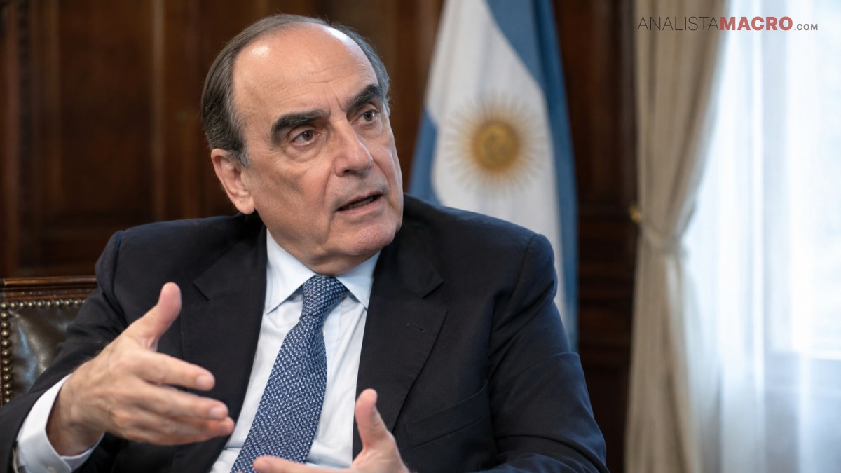 Hombre de traje oscuro y corbata azul gesticula sentado, con una bandera argentina y cortinas claras al fondo.