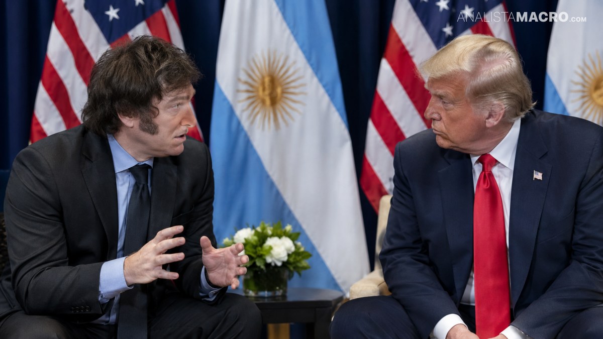 Dos hombres conversan sentados frente a banderas de Estados Unidos y Argentina, con un pequeño arreglo floral entre ambos.