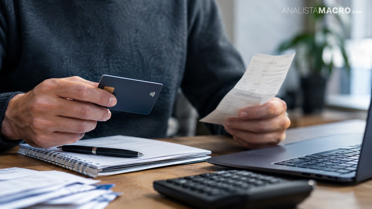 Persona sostiene una tarjeta bancaria y un recibo frente a un cuaderno, una calculadora y una computadora portátil.