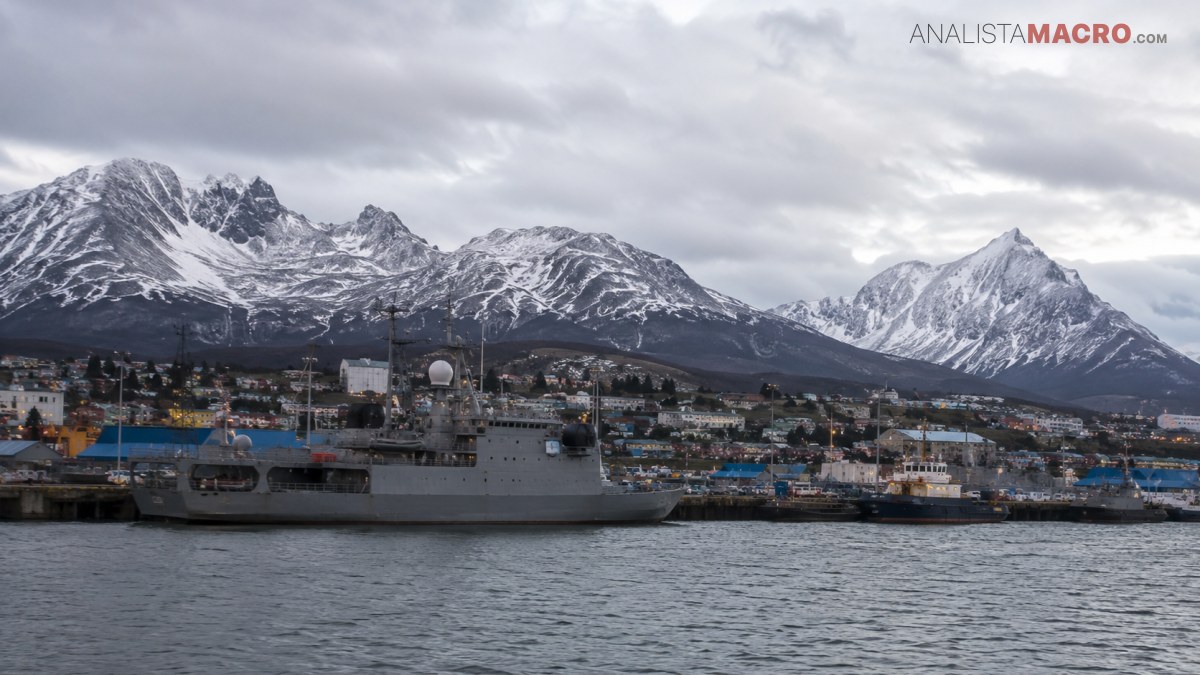Buque gris amarrado frente a una ciudad costera, con montañas nevadas y cielo nublado al fondo.
