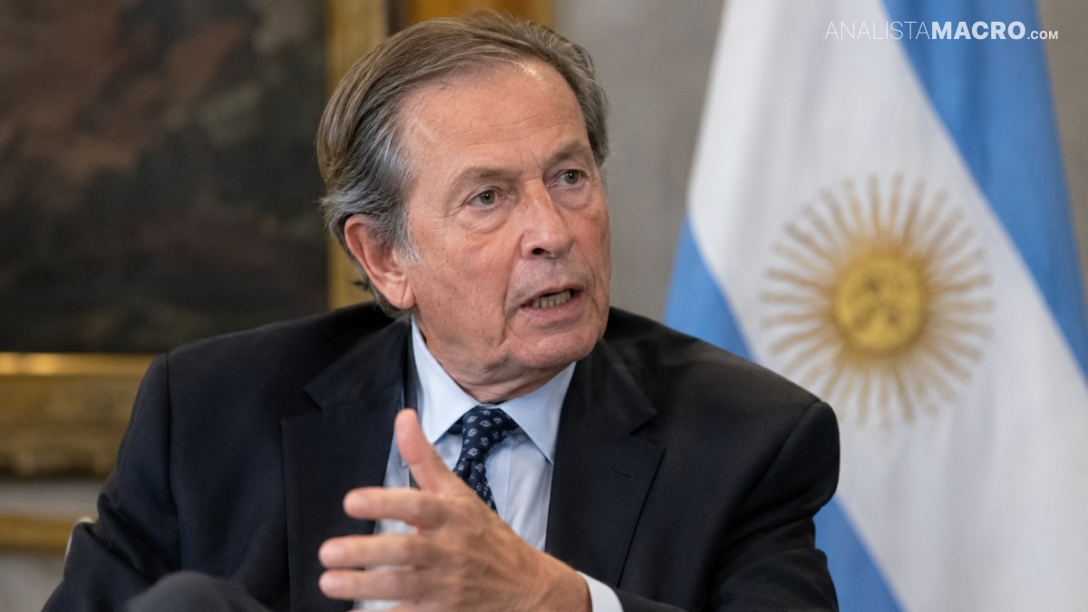 Hombre de cabello canoso con traje oscuro gesticula mientras habla, con una bandera argentina visible al fondo.
