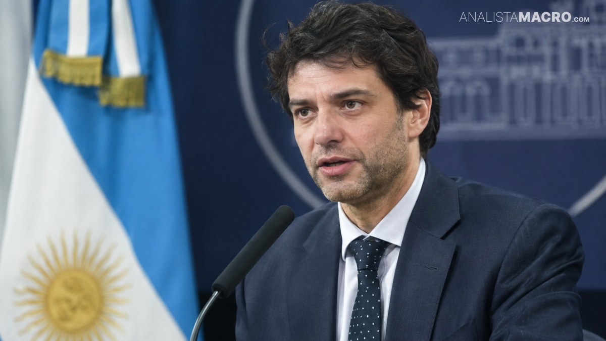 Hombre de traje oscuro y corbata hablando ante un micrófono, con una bandera argentina y un fondo institucional.