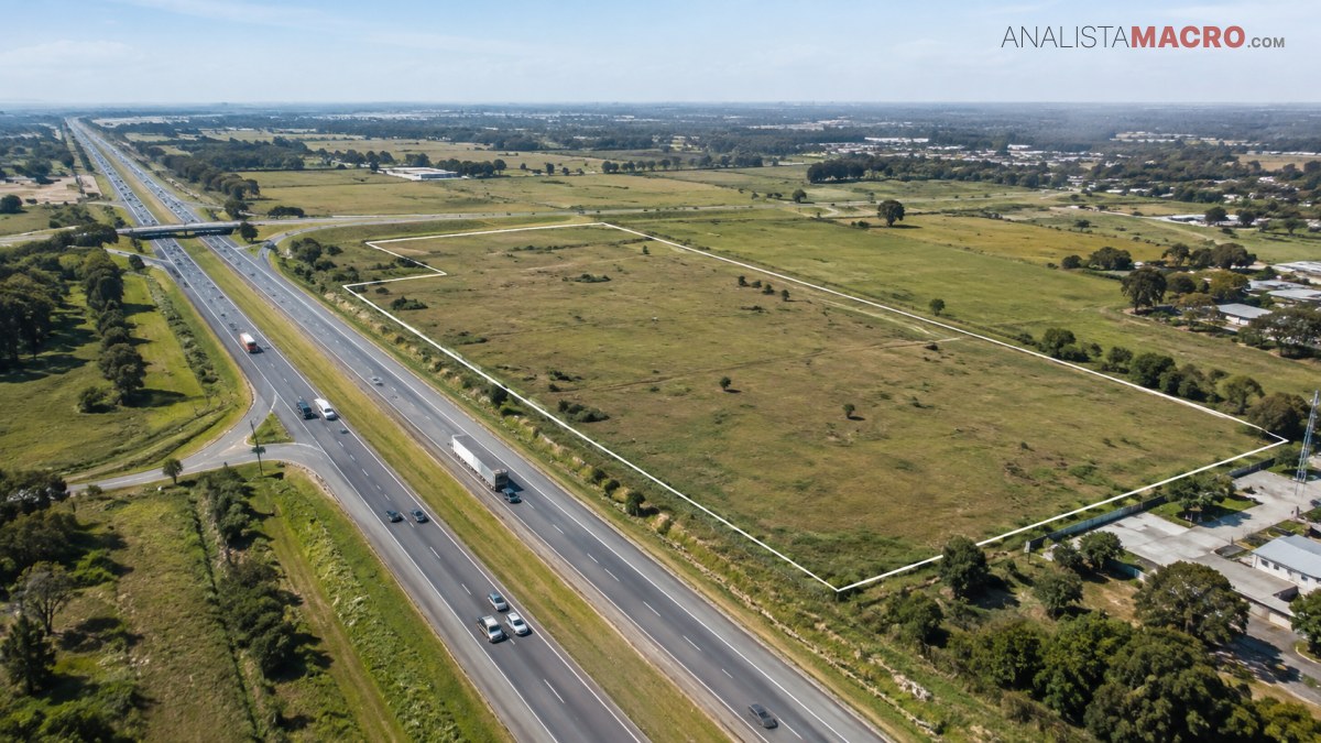 Autopista de varios carriles junto a extensos terrenos verdes, viviendas dispersas y un paisaje urbano visible en el horizonte.