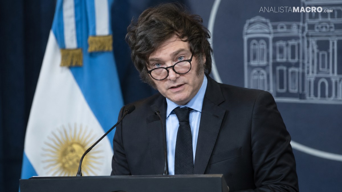 Persona con gafas y traje negro habla en un atril con dos micrófonos, con bandera y fondo arquitectónico detrás.