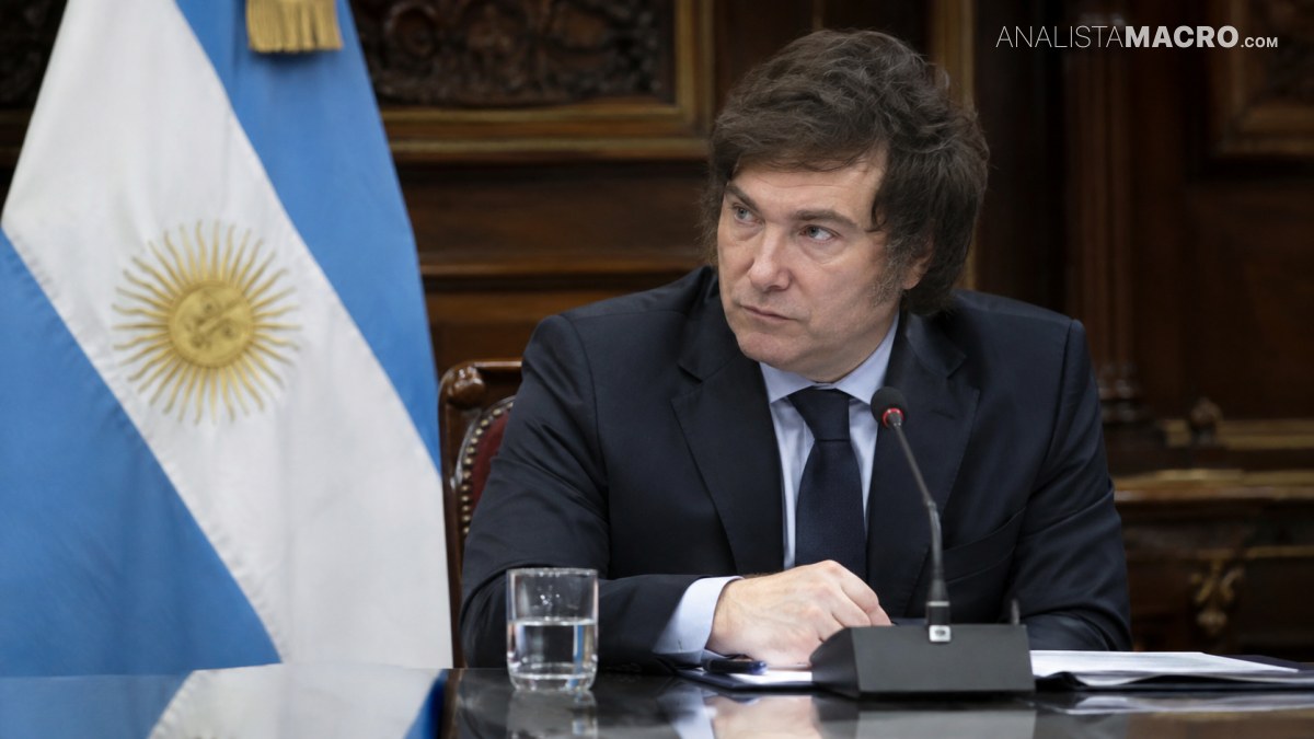 Hombre de traje oscuro sentado ante un micrófono, con una bandera argentina y documentos sobre la mesa.