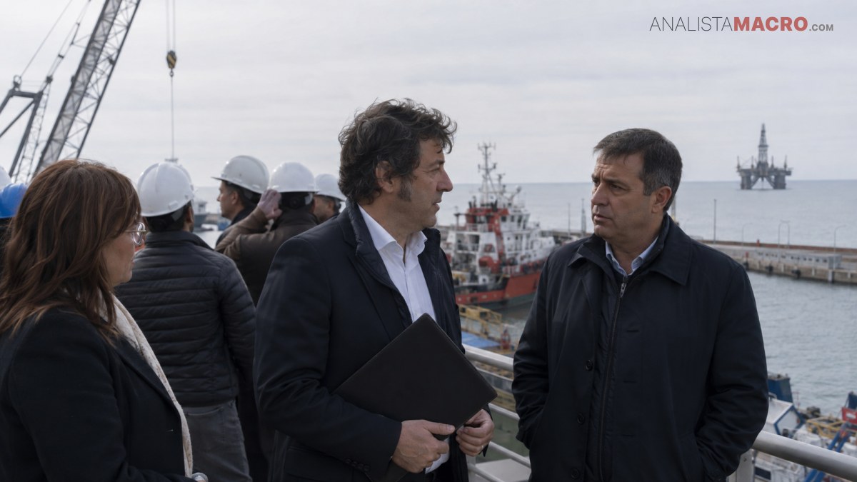 Tres personas conversan frente a un puerto, mientras trabajadores con cascos, grúas, barcos y plataformas marinas aparecen al fondo.