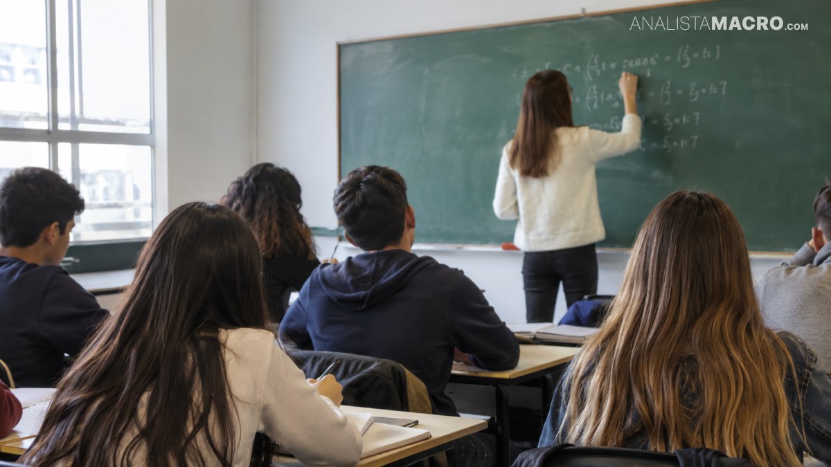 Estudiantes sentados en un aula observan a una profesora escribir fórmulas en una pizarra.