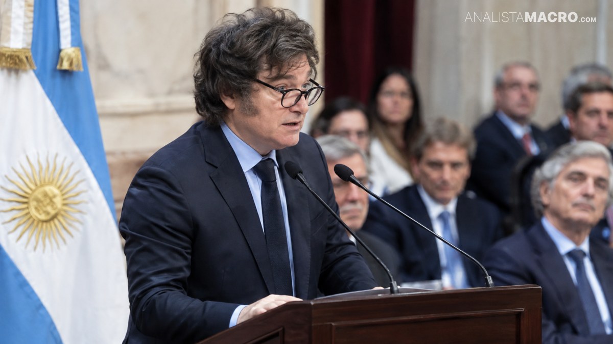 Hombre con gafas y traje oscuro habla ante un micrófono, con una bandera argentina y varias personas sentadas detrás.