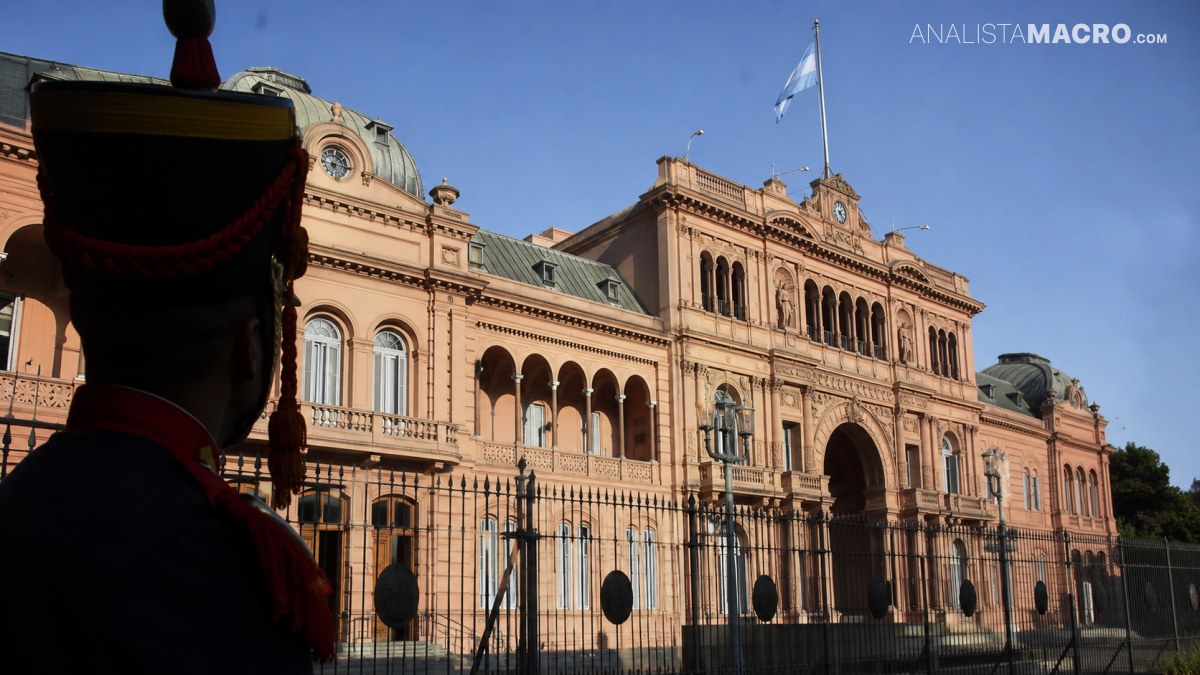 Edificio histórico de fachada rosada con cúpulas y banderas, rodeado por una reja y parcialmente oculto por un guardia uniformado.