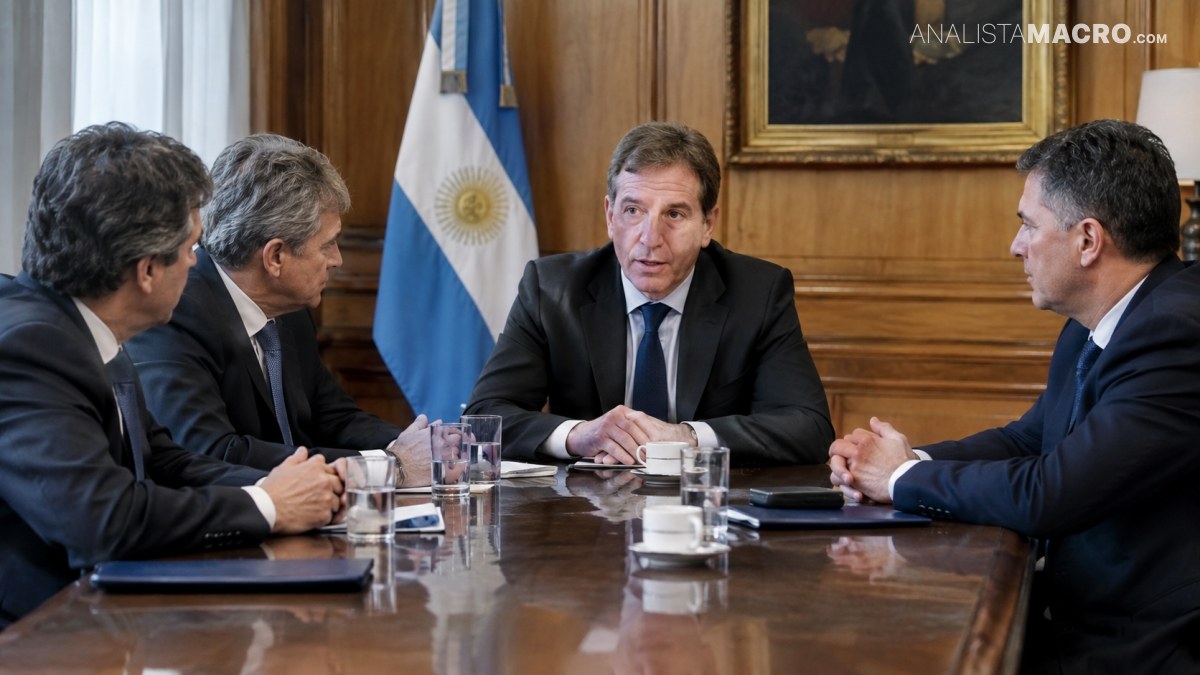 Cuatro hombres con trajes conversan alrededor de una mesa, junto a una bandera argentina y vasos de agua.