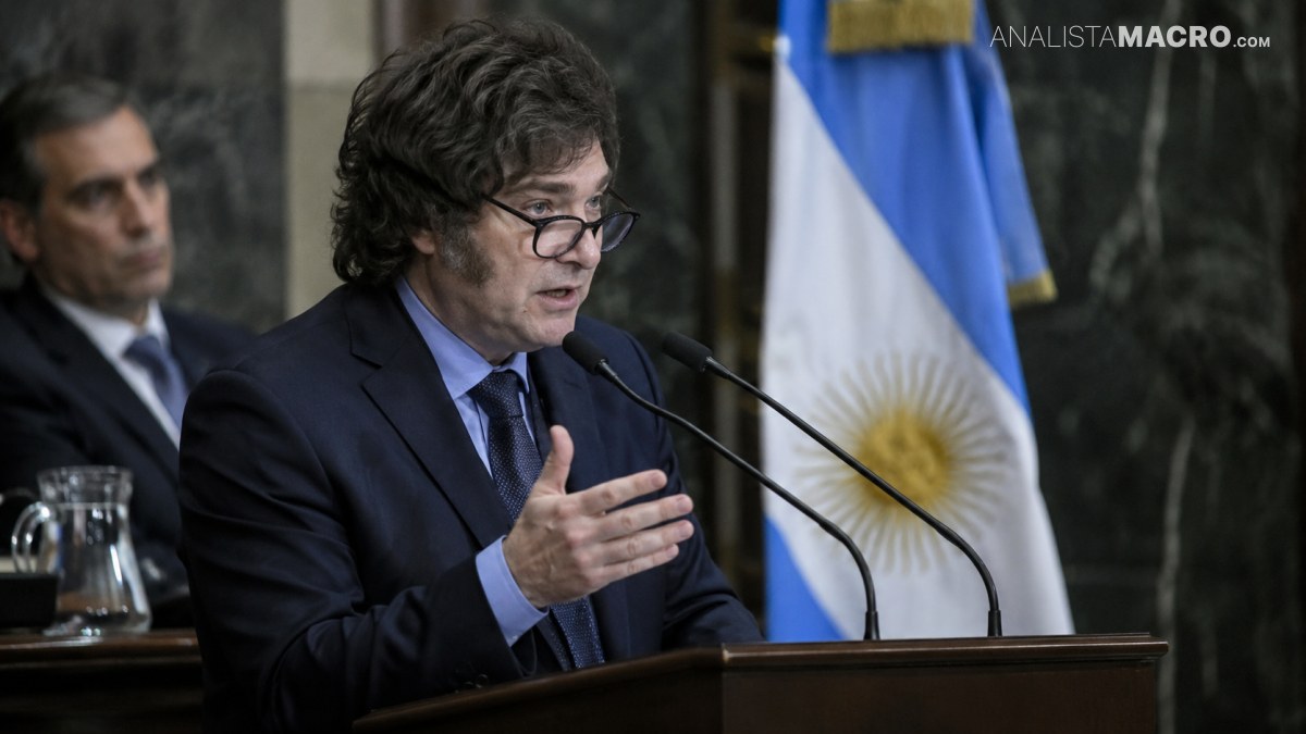 Hombre con gafas habla ante dos micrófonos, gesticulando con una mano, mientras una bandera celeste y blanca aparece detrás.