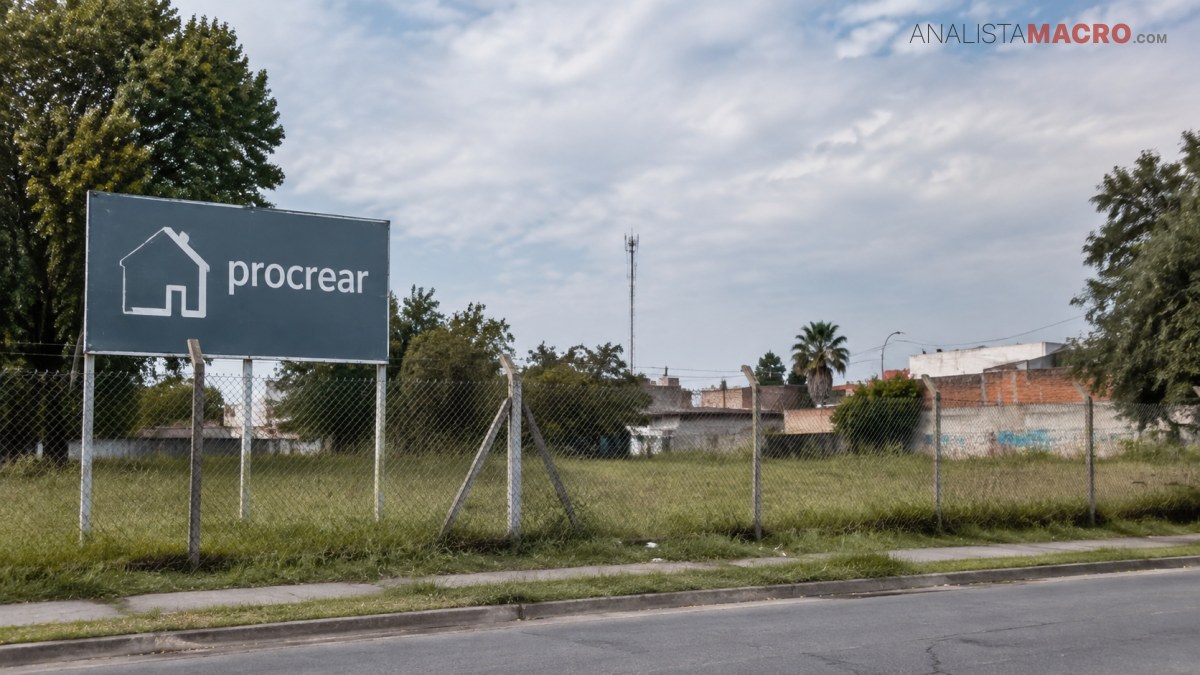 Cartel de Procrear junto a un terreno cercado, con viviendas, árboles y una calle asfaltada bajo un cielo nublado.