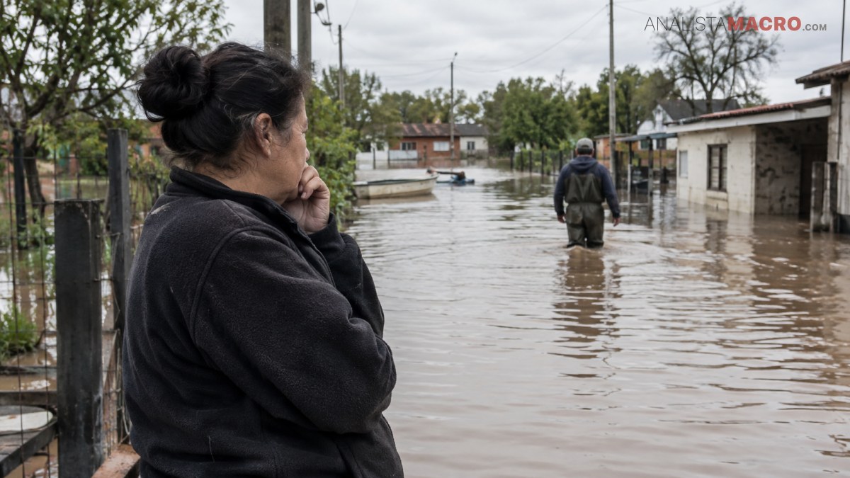 Mujer observa a una persona caminando por una calle inundada, rodeada de viviendas, árboles y postes de electricidad.