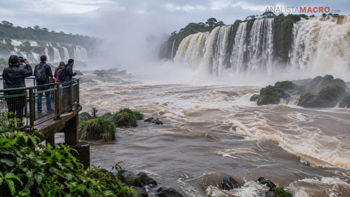 Personas observan una amplia cascada desde una plataforma metálica, rodeada de vegetación, rocas y agua turbulenta.