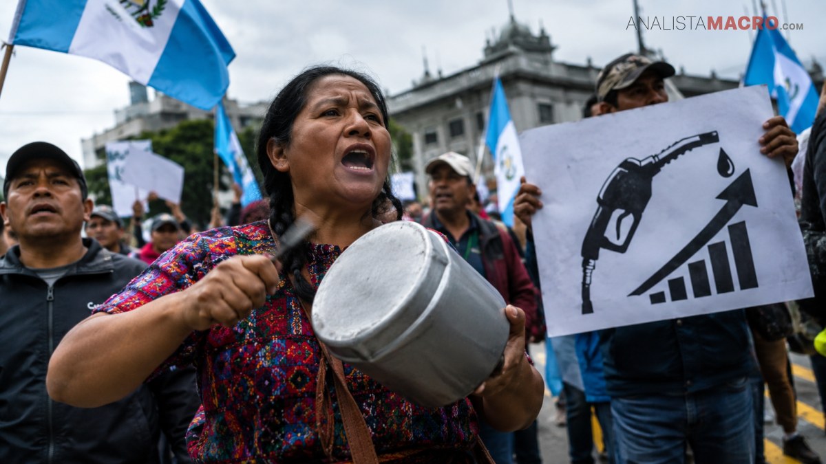 Mujer con vestimenta colorida golpea un recipiente metálico durante una protesta, mientras otros sostienen banderas y un cartel sobre combustible.