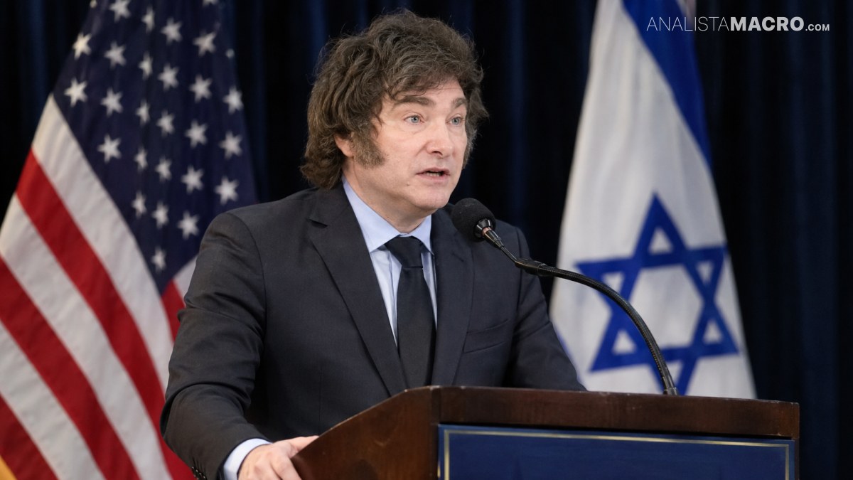 Hombre de cabello despeinado habla ante un micrófono desde un atril, con las banderas de Estados Unidos e Israel al fondo.