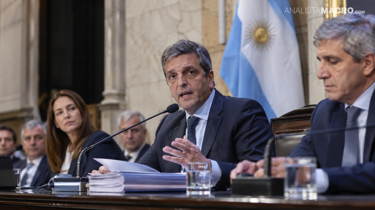 Hombre de traje habla ante un micrófono, sentado junto a otros funcionarios, con una bandera argentina al fondo.