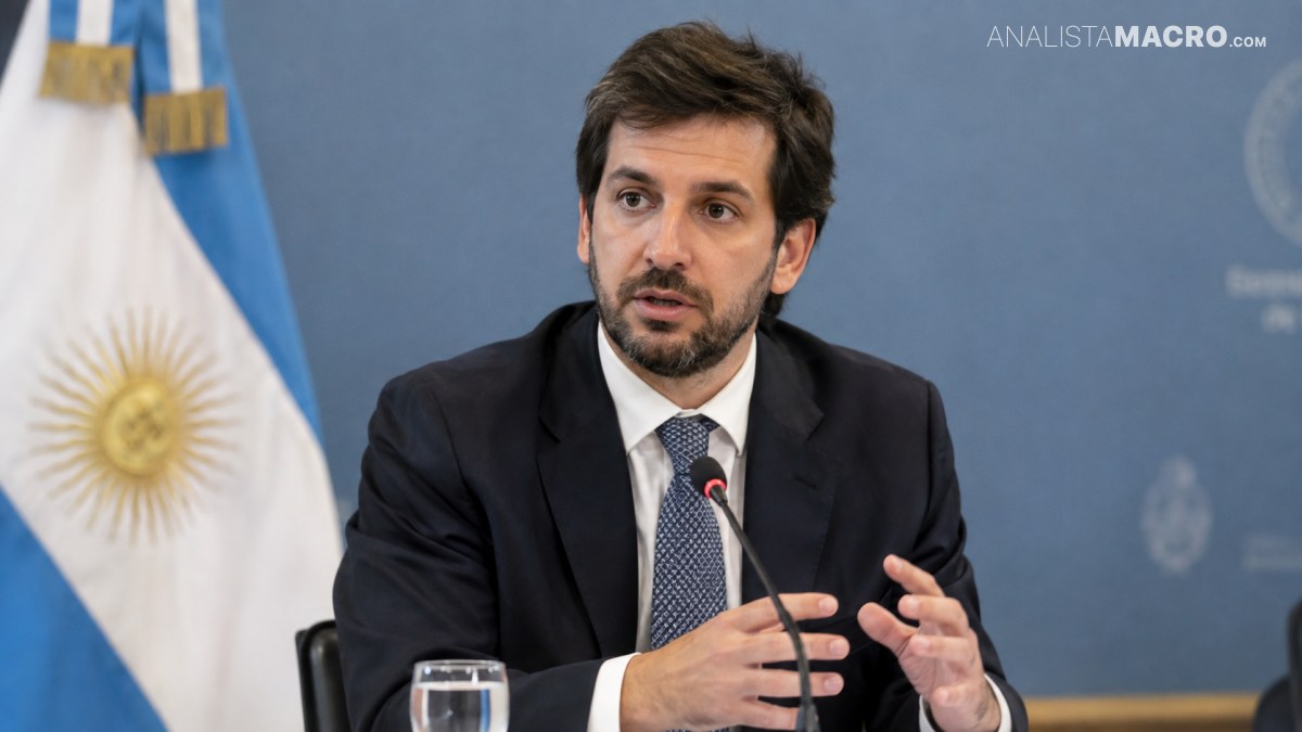 Hombre con traje oscuro habla ante un micrófono, sentado frente a una bandera argentina y un fondo azul institucional.