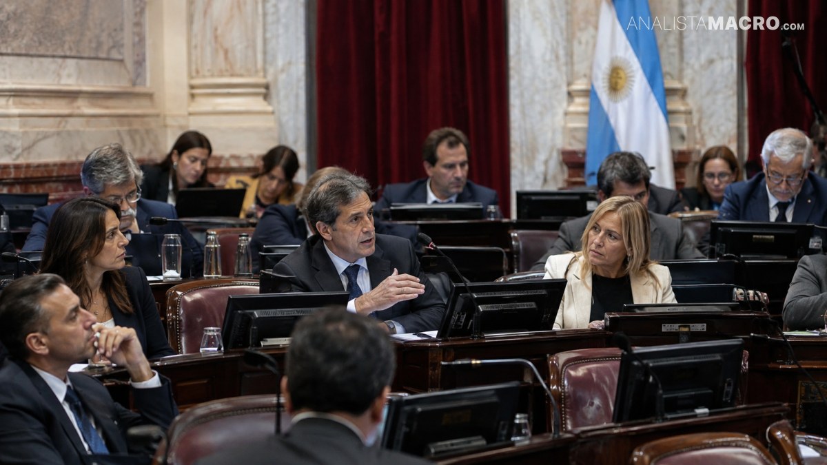 Personas sentadas en un recinto formal, conversando entre escritorios con pantallas, mientras una bandera argentina aparece al fondo.